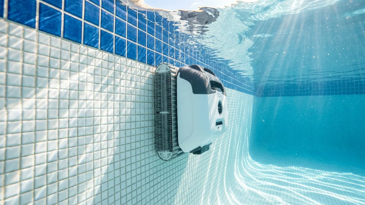 Vue sous l'eau d'un robot de piscine qui monte le long d'une paroi carrelée de mosaïque blanche vers la ligne d'eau bleue
